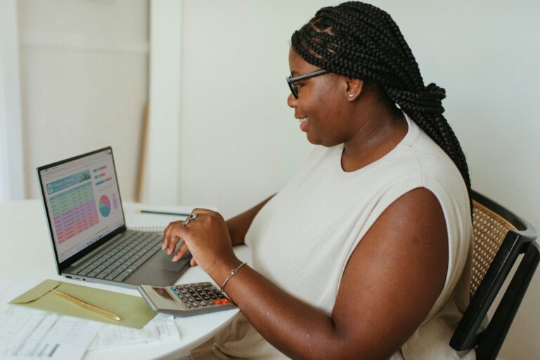 A Black women with long hair and glasses is looking at a computer that is displaying analytics on the screen.